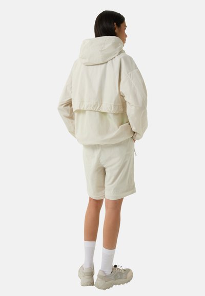 Light beige cropped jacket with hood, layered design, elastic cuffs, paired with matching shorts, and white socks. Textured fabric, casual fit.