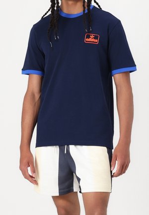 Personne portant un t-shirt Adidas bleu marine avec un liseré bleu et un short bicolore noir et blanc sur un fond blanc uni.