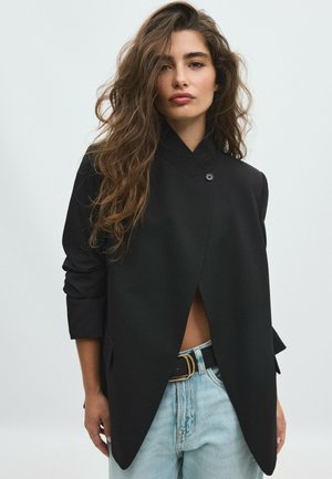 Frau mit langem, welligem Haar trägt eine schwarze asymmetrische Jacke mit Knöpfen über hellblauen Jeans mit schwarzem Gürtel, stehend vor einem einfarbigen Hintergrund.
