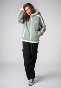 Groene fleece jas met witte accenten, ritssluiting zakken en een capuchon, gecombineerd met zwarte cargobroeken en zwart-witte sneakers.