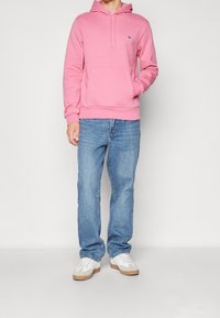 Hoodie rosa in un morbido tessuto con tasca centrale davanti e cappuccio con lacci, abbinato a jeans azzurri chiari e sneakers bianche.