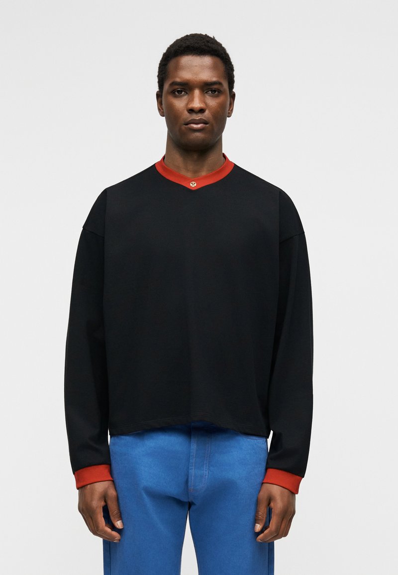 Homme portant une chemise noire à manches longues avec col et poignets rouges, associée à un pantalon bleu, debout devant un fond clair uni.