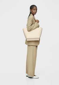 Beżowa torba na ramię o gładkiej fakturze, z wypukłym logo marki. Projekt jest sztywny, z szerokim otwarciem i kontrastowym dnem.