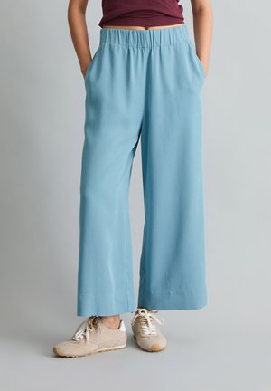 Persoon die lichtblauwe wijduitlopende broek met elastische tailleband en beige sneakers draagt, staand tegen een neutrale achtergrond.