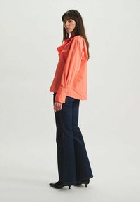 Chemise corail à manches larges, associée à un jean évasé bleu foncé et des bottes à talons noires. Tissu lisse, détails de boutons aux poignets.