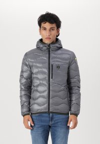 Graue Steppjacke mit Kapuze, die über gewellte Nähte, einen Reißverschluss auf der Vorderseite und eine Brusttasche verfügt. Glänzendes, strukturiertes Material mit grünen Akzenten.