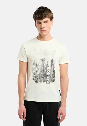 Ung mand iført en hvid T-shirt med en sort og grå bysilhuet-grafik og sorte bukser, stående mod en ensartet baggrund.