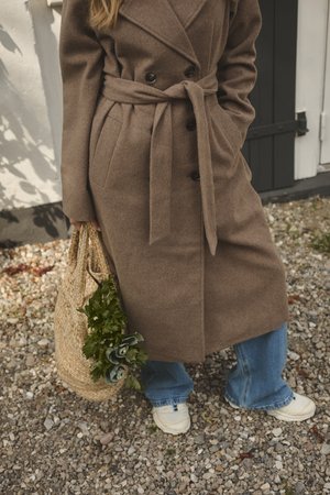 Brauner wollener Doppelreiher-Mantel mit Gürtel, kombiniert mit blauen Jeans und weißen Sneakers. Hält eine Stroh-Tasche mit grünen Kräutern.