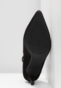 Chaussure noire à talon haut et bout pointu, posée sur le talon, semelle visible avec motif antidérapant texturé et indication de taille.