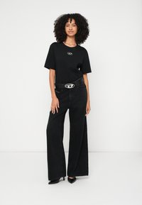 Γυναίκα με σγουρά μαλλιά που φοράει μαύρο μπλουζάκι, μαύρο τζιν παντελόνι με φαρδιά μπατζάκια, ζώνη με ασημένια αγκράφα και μαύρες μυτερές γόβες, πάνω σε λευκό φόντο.