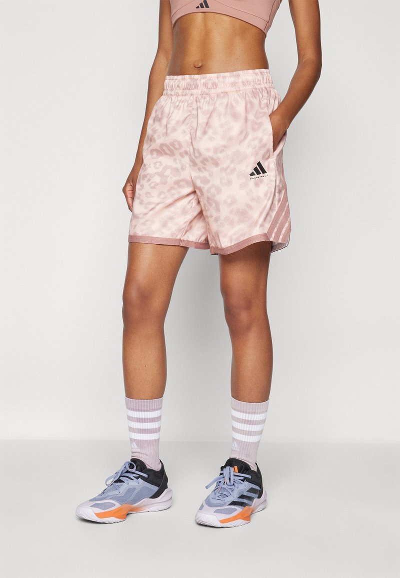 Femme portant un short Adidas rose à motifs, des chaussettes rayées et des chaussures de sport grises, debout avec une main dans la poche.