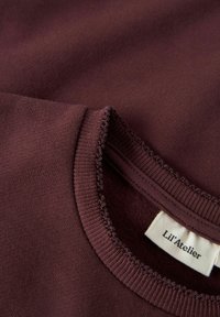 Detailní záběr na tmavě vínovou látku z fleecu, na kterém je vidět jemné šití výstřihu a bílá etiketa s názvem značky Lil'Atelier.