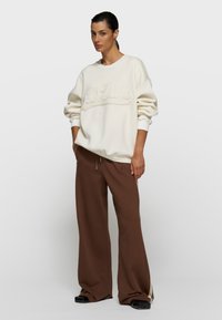 Sudadera blanca con letras en relieve "10 Days", combinada con pantalones deportivos marrones de corte ancho con franjas laterales y cordón ajustable.