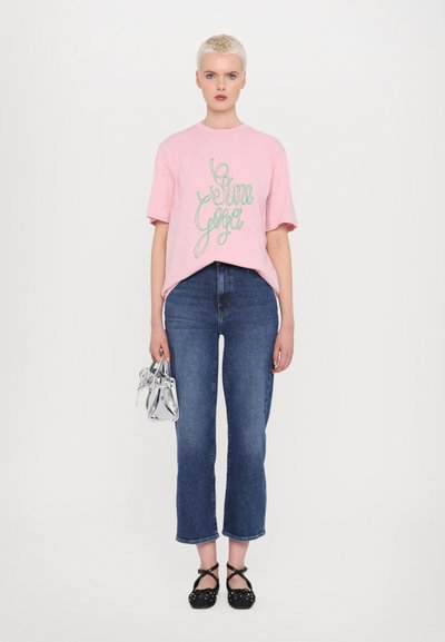 Pink cotton t-shirt with green lettering, paired with high-waisted blue jeans. Model holds a small clear handbag and wears black dotted shoes.