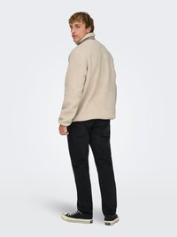 Veste en polaire beige avec un col montant, associée à un jean noir et des baskets noires avec des accents blancs. Tissu texturé, coupe décontractée.