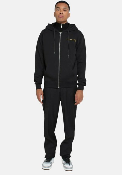 Felpa nera con zip frontale, dotata di un logo dorato. Abbinata a pantaloni neri e sneakers bianche e blu. Tessuto liscio, vestibilità informale.