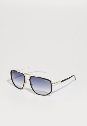 Lunettes de soleil avec une monture en métal doré, verres carrés dégradés du gris foncé au clair. Elles possèdent des branches en plastique blanc et noir avec des accents de logo.