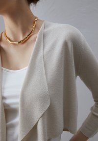 Žena v bílé tílku, béžovém pleteném kardiganu a masivním lesklém zlatém náhrdelníku, na neutrálním pozadí.