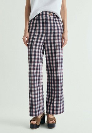 Pantalones de pierna ancha a cuadros con detalle de botones delanteros, combinados con top blanco y sandalias marrones de plataforma sobre un fondo claro.