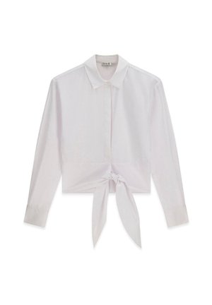 Jeune femme debout en plein air portant une chemise blanche nouée, un pantalon capri bleu à motifs et des sandales blanches à enfiler, posant pour des photos.