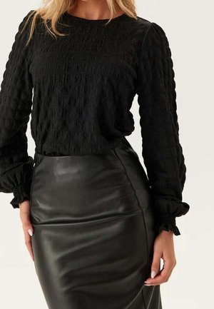 Femme portant un haut noir texturé à manches longues et une jupe en cuir noire taille haute, debout les mains le long du corps.