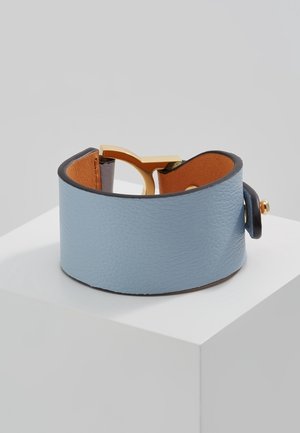 Bred lyseblå læderarmbånd med tanfarvet inderside og guldfarvet spænde, vist på hvid piedestal mod grå baggrund.