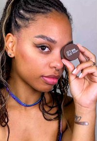 Braune Creme-Dose mit der Bezeichnung "575", die neben dem Gesicht gehalten wird, mit matter Oberfläche. Mehrfarbige Perlenkette und dezenter Lippenstift sind sichtbar.