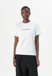 T-shirt de algodón blanco con mangas cortas, cuello redondo acanalado y "Calvin Klein" impreso en negro en el pecho. Textura suave.