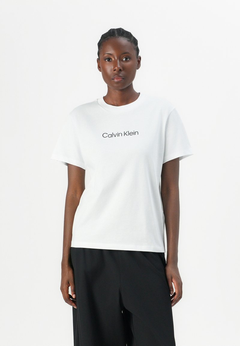 T-shirt de algodón blanco con mangas cortas, cuello redondo acanalado y "Calvin Klein" impreso en negro en el pecho. Textura suave.