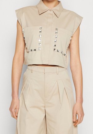 Beige cropped sleeveless shirt with structured shoulders and vertical jewel embellishments, paired with high-waisted pleated beige pants.
