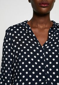 Blusa navy con pois bianchi, colletto e chiusura con bottoni davanti. Presenta maniche lunghe e una texture di tessuto liscia e leggera.