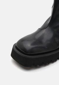 Stivale alla caviglia in pelle nera con punta rotonda, suola spessa e robusta, e texture liscia. Presenta una leggera lucentezza e nessun hardware visibile.