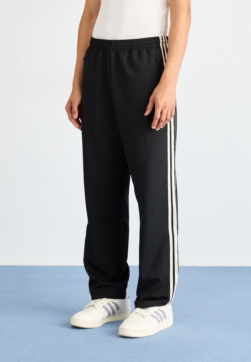 Černé sportovní kalhoty pohodlného střihu, se zvýrazněnými bílými bočními pruhy, elastickým pasem a kapsami na zip. V kombinaci s bílými teniskami.