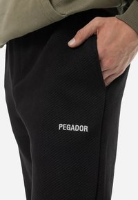 Pantalones jogger negros con relieve, con bolsillos laterales y un logo blanco "PEGADOR" en el muslo izquierdo. Presentan un ajuste relajado y una tela texturizada.