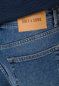 Blå denimjeans med et lærmerke som leser "ONLY & SONS." Har synlig søm, lommer og en teksturert stofffinish.