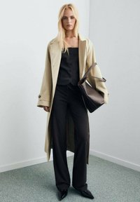 Trench coat beige su una blusa nera con bottoni e pantaloni neri a gamba larga, abbinato a scarpe nere appuntite e una borsa marrone.