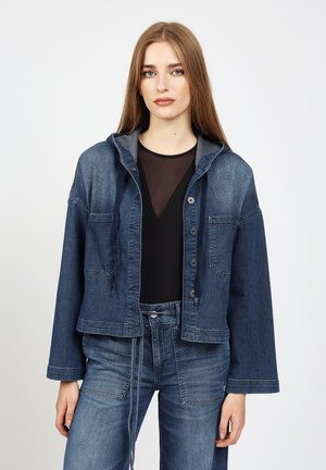 Junge Frau mit langen Haaren, die eine blaue Jeansjacke mit großen Taschen und passende Jeans vor einem einfarbigen Hintergrund trägt.