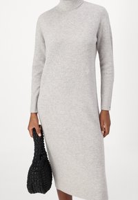 Robe en maille côtelée grise avec un col montant et des manches longues, associée à un sac à main tissé noir texturé. Modèle mi-long.