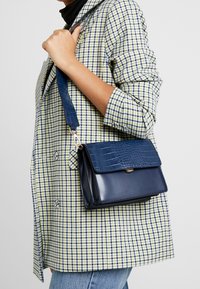 Borsa a tracolla blu navy con texture a coccodrillo sulla patta, corpo liscio e tracolla a quadri; presenta una chiusura magnetica.