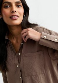 Bruine, gekreukelde stoffen shirt met een kraag, knoopsluiting aan de voorkant en een borstzak. Beschikt over lichte knopen en lange mouwen met knoopmanchetten.