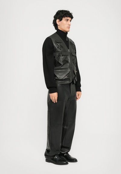 Man wearing black leather utility vest with multiple pockets, black turtleneck sweater, loose black leather pants, and black dress shoes standing sideways.