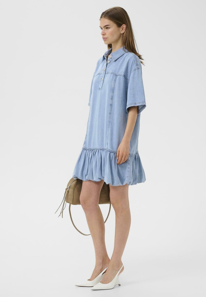 Vrouw staand, draagt een losse lichtblauwe denim jurk met korte mouwen en een gerufflede zoom, houdt een beige handtas vast en witte hakken.