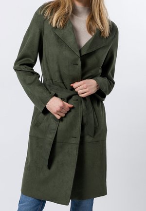 Femme portant un manteau ceinturé vert olive mi-long sur un col roulé beige et un jean bleu, en train d'attacher la ceinture du manteau.