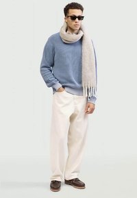 Junger Mann mit schwarzen Sonnenbrillen, blauer gerippter Pullover, beigem Schal, weißen Hosen und braunen Schuhen, der mit einer Hand in der Tasche steht.