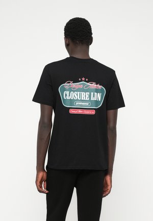 Juodas marškinėlis su grafiniu spaudiniu ant nugaros, su spalvingu tekstu ir žvaigždėmis. Medžiaga atrodo minkšta ir pralaidi orui.