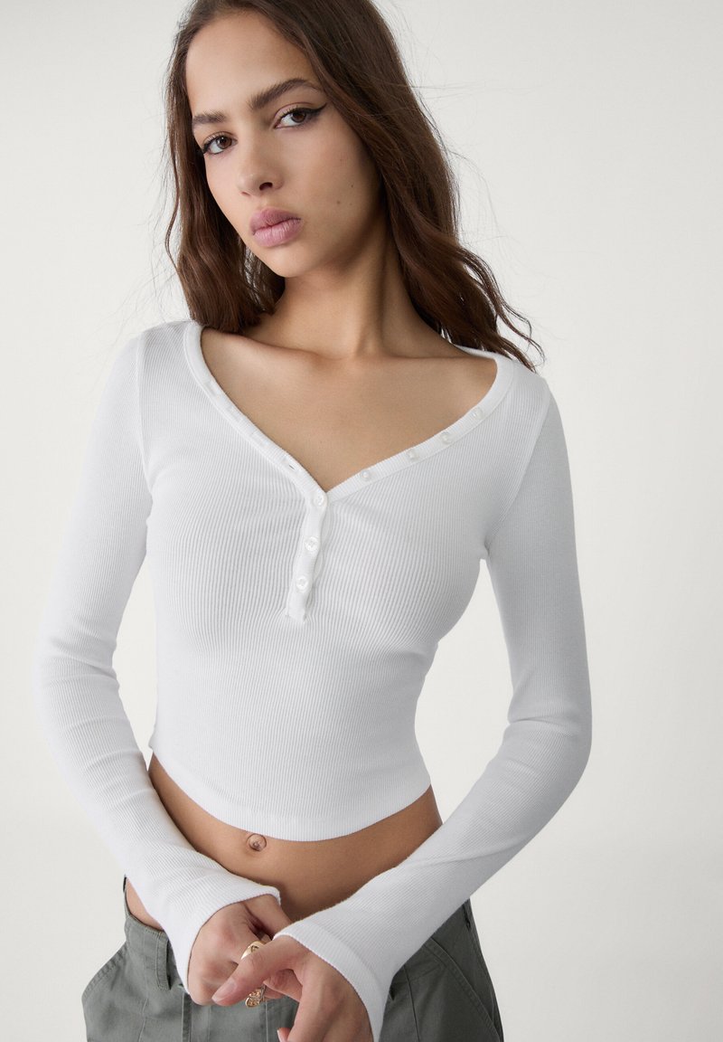 White ribbed long-sleeve top with a V-neck, featuring buttons along the neckline and fitted design. The bottom hem is cropped, exposing the midriff.