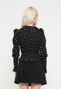 Bluza neagră cu buline cu detalii smocate și mâneci bufante. Spatele are un design adunat, asortat cu o fustă neagră.