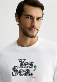Hvid bomulds-T-shirt med mørke paisley-mønstrede bogstaver, der står "Yes, Sea." samt et rødt "LIU-JO" logo nederst til højre.