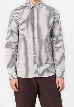 Person wearing a long-sleeve, buttoned shirt with vertical navy and white stripes, paired with dark brown pants, standing against a plain background.