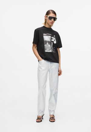 Ženska v črni majici z grafičnim potiskom, svetlo modrih kavbojkah, črnih sandalih s peto in velikih belih sončnih očalih stoji z roko v žepu.